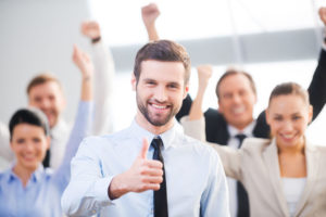 34391345 - feeling confident in his team. happy businessman showing his thumb up and smiling while his colleagues standing in the background